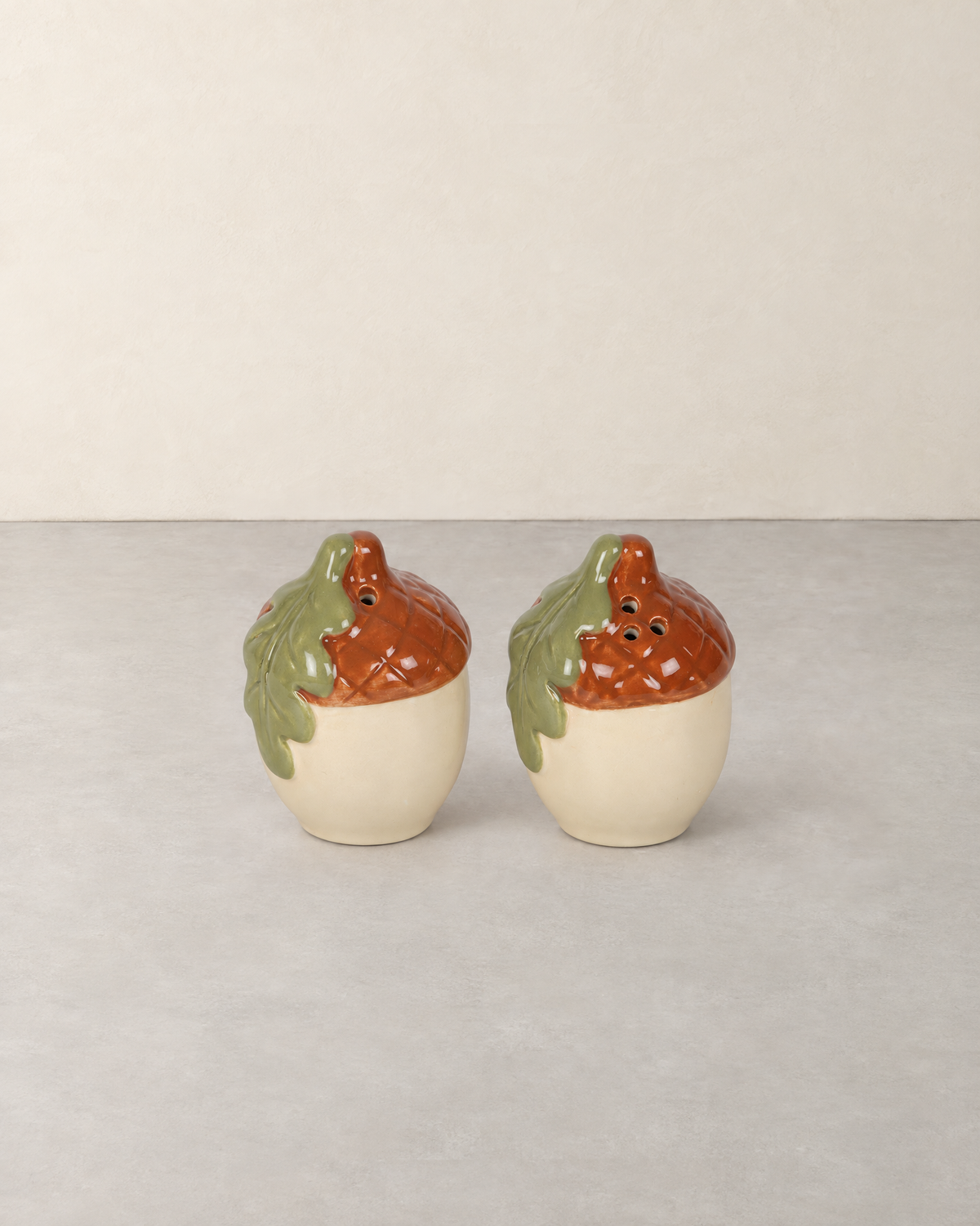 Pair of ceramic acorn salt and pepper shakers with russet caps and green oak leaves on a pale grey floor against a cream wall