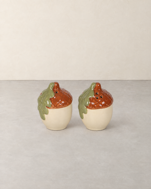 Pair of ceramic acorn salt and pepper shakers with russet caps and green oak leaves on a pale grey floor against a cream wall