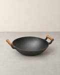 Black cast iron wok with two wooden handles, standing empty on a pale concrete floor