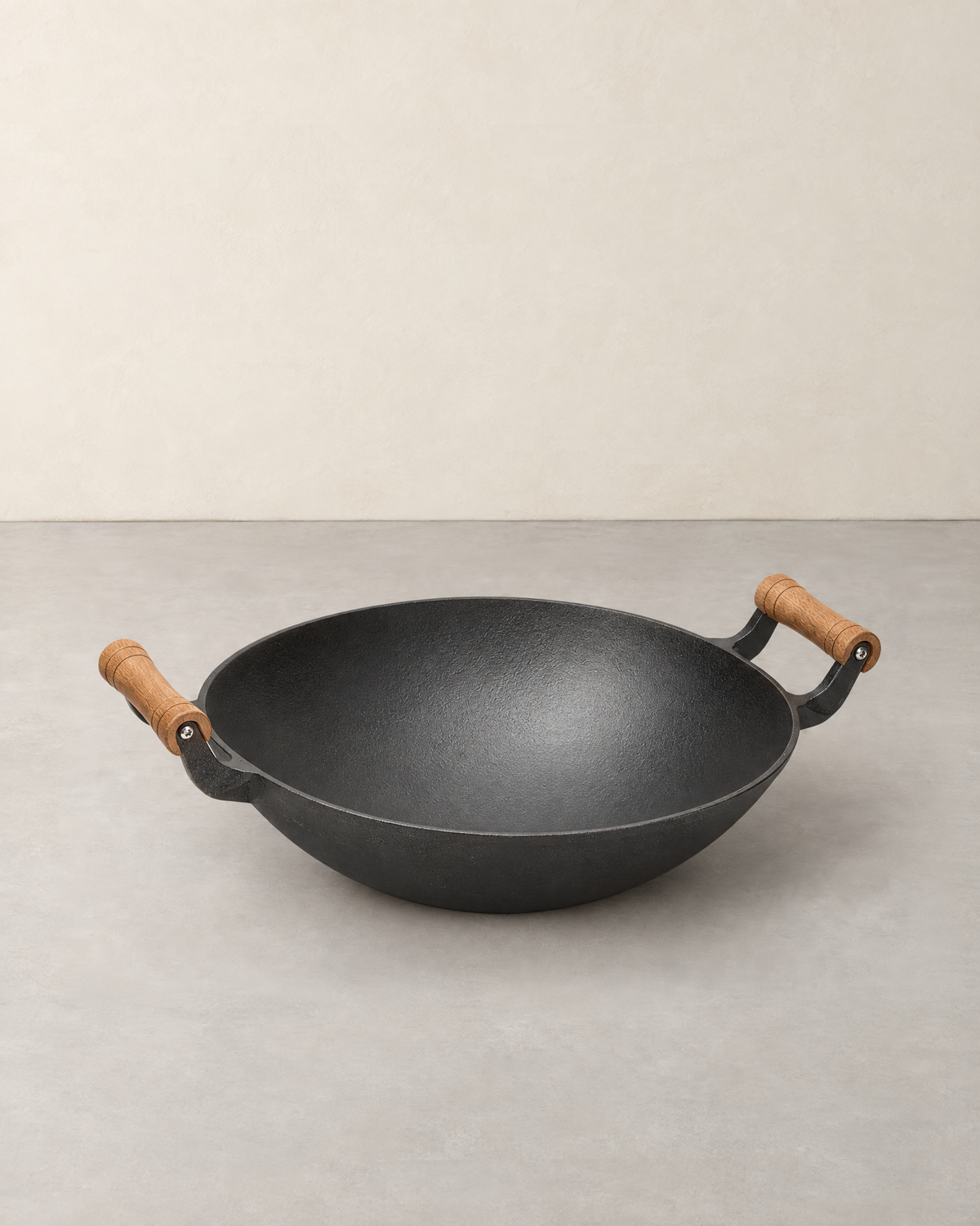 Black cast iron wok with two wooden handles, standing empty on a pale concrete floor