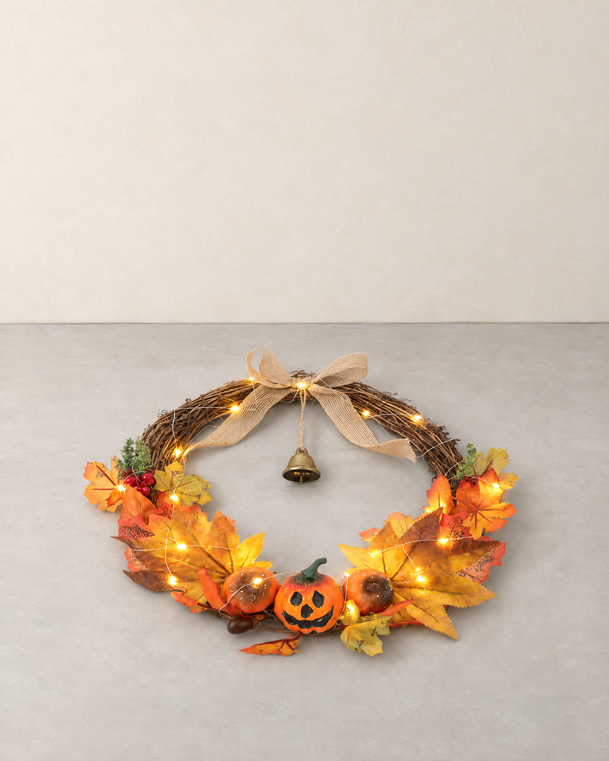 Open woven branch harvest wreath with maple leaves, carved-face pumpkins, a burlap bow and a bell, lying flat