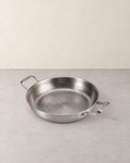 Hammered titanium skillet with two side handles standing empty on a pale concrete floor against a cream wall
