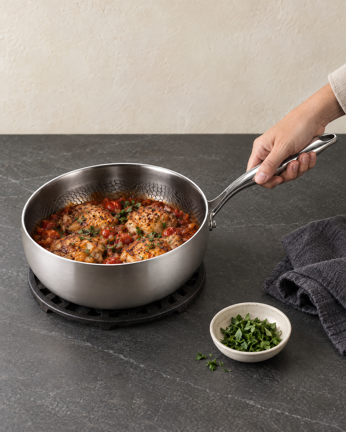 The titanium saute pan with chicken braised in tomato sauce on a trivet, a hand holding its long handle