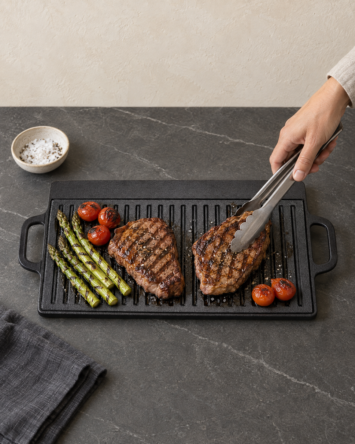 Steaks, asparagus and cherry tomatoes cooking on the ribbed cast iron griddle, turned with steel tongs