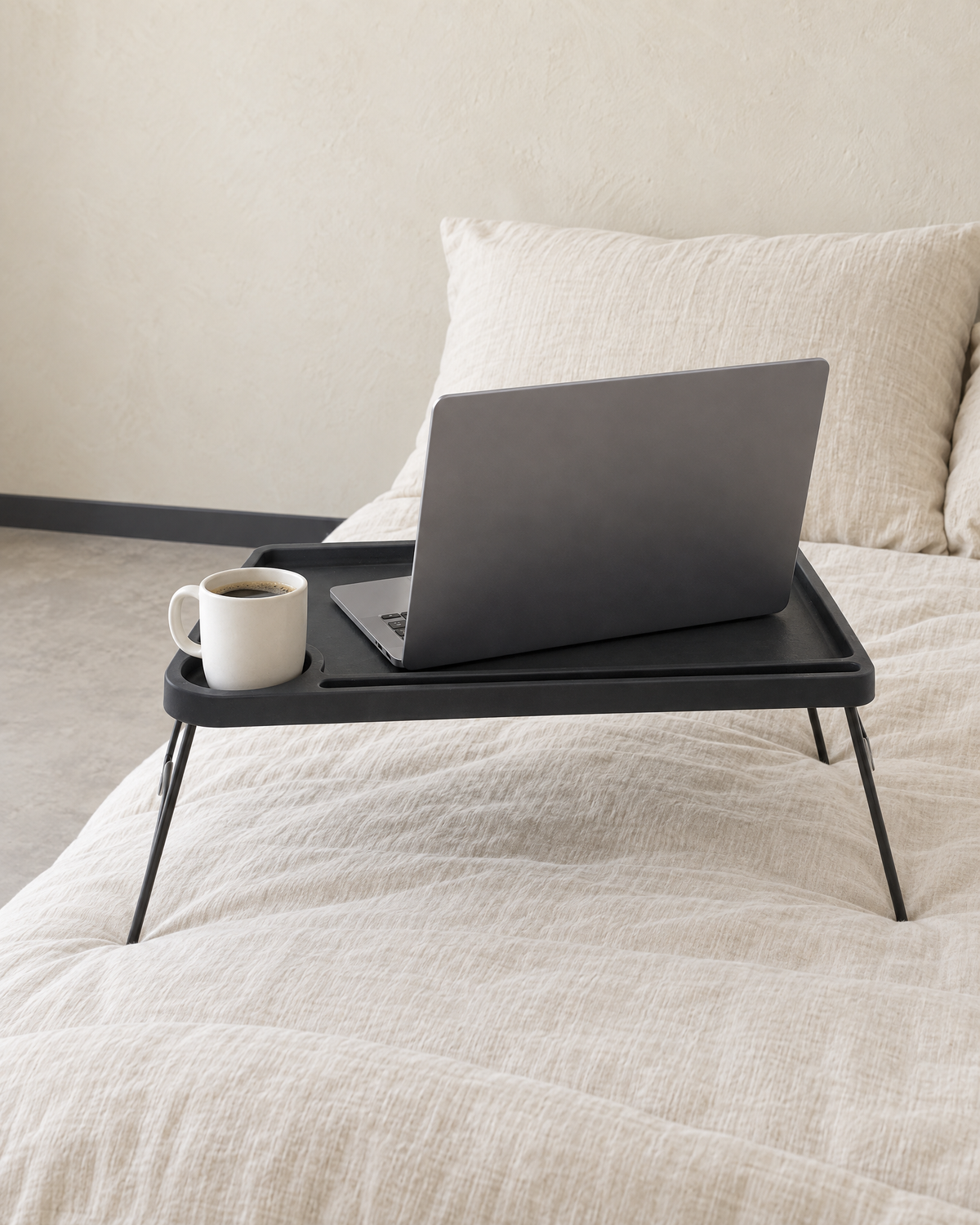 The black folding bed table on a bed holding an open laptop and a mug in the cup holder