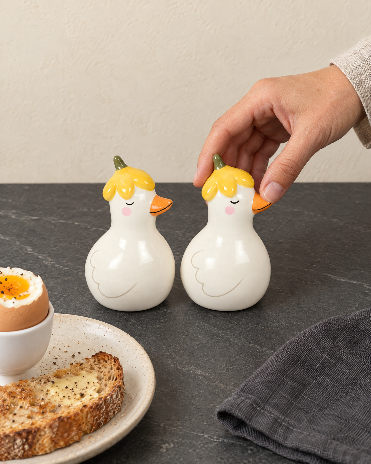 Two duck shakers in yellow flower hats beside a boiled egg and toast, one about to be picked up