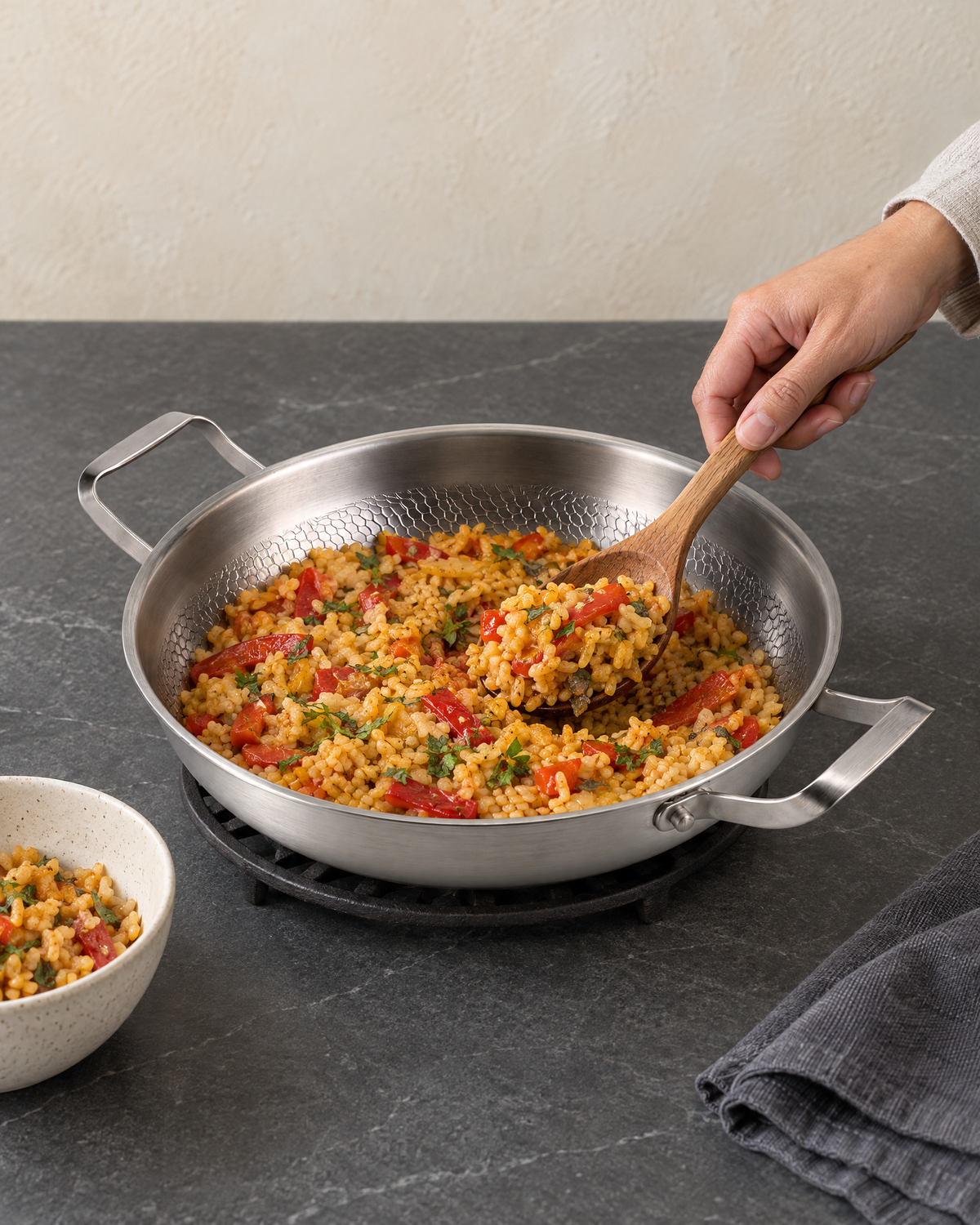 The two-handled titanium skillet full of a golden rice dish on a trivet on a dark stone worktop, a hand serving with a wooden spoon