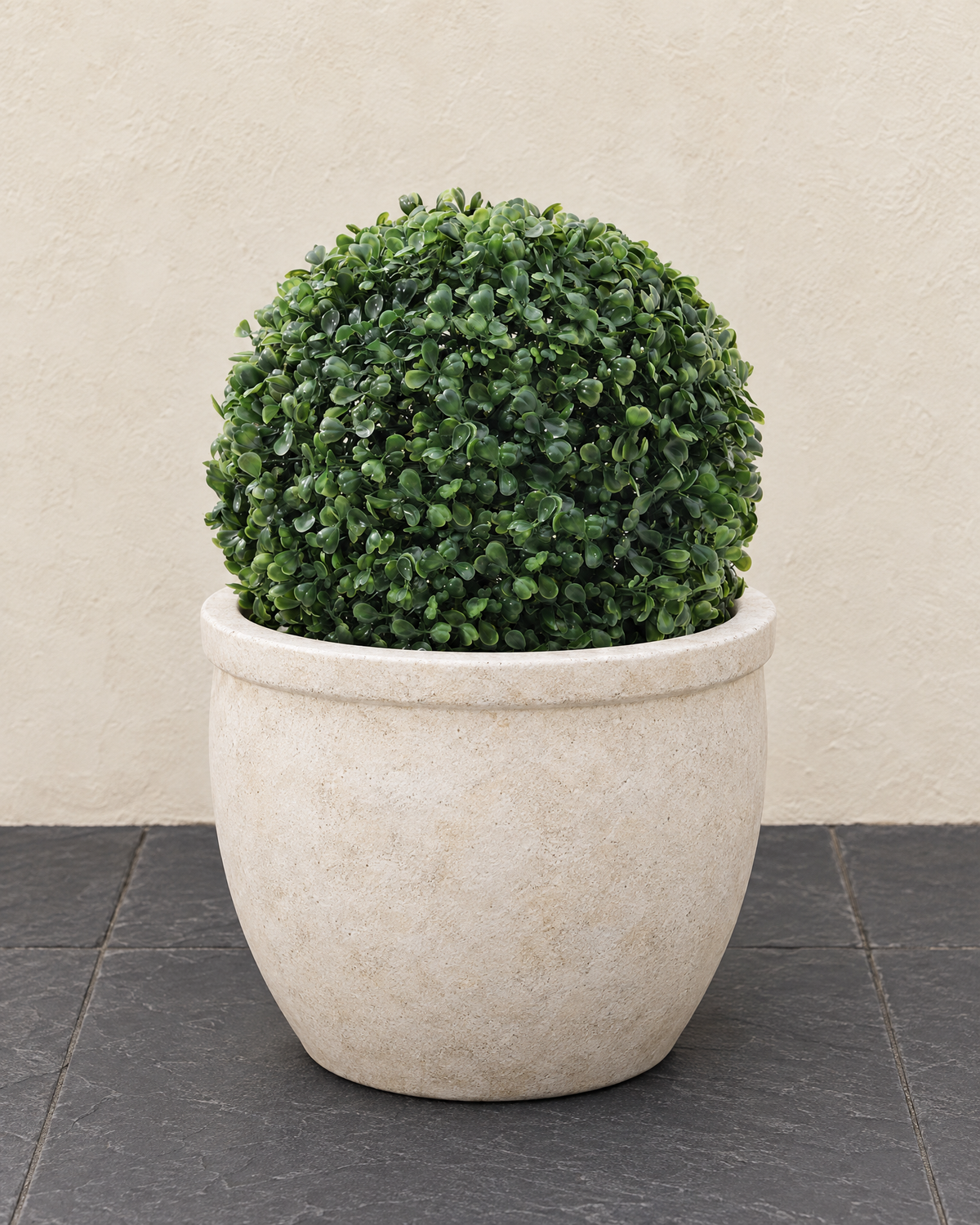 Artificial boxwood ball sitting in a round cream stone planter on charcoal slate paving by a cream garden wall
