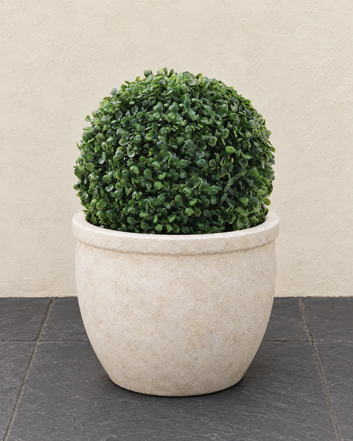 Artificial boxwood ball sitting in a round cream stone planter on charcoal slate paving by a cream garden wall