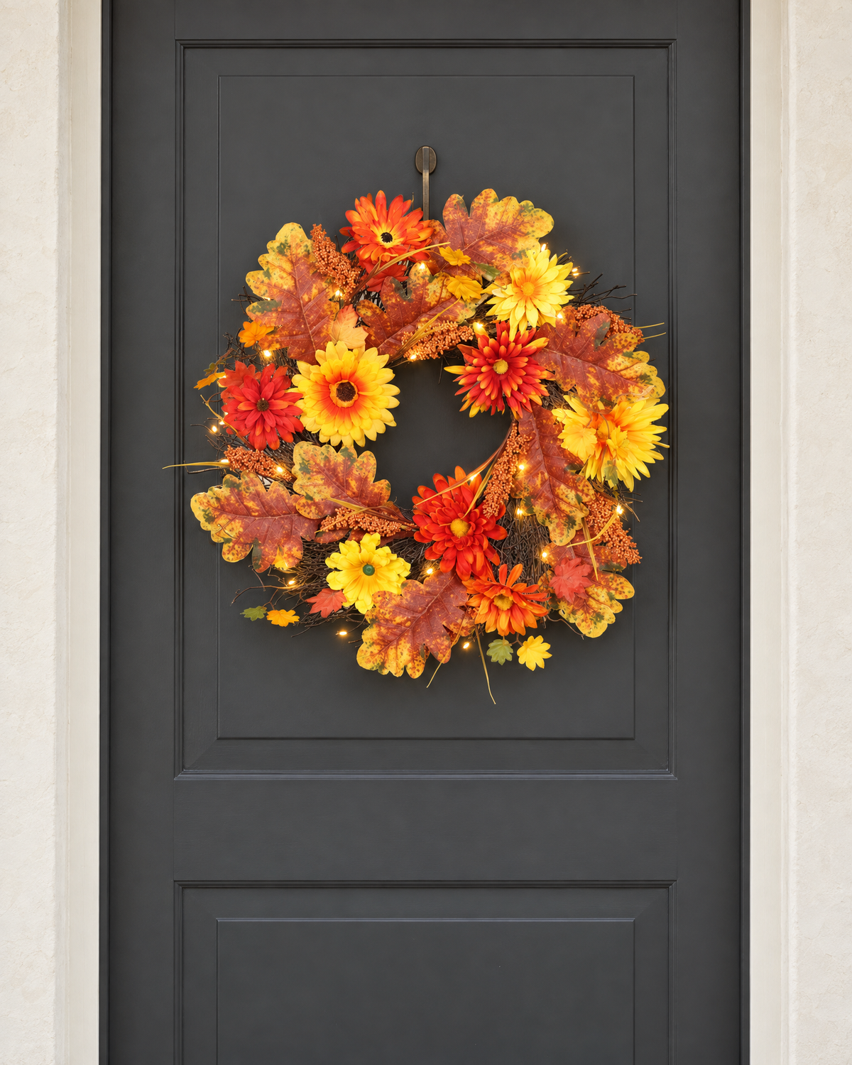 The daisy and maple leaf wreath with its lights on, hung on a charcoal front door