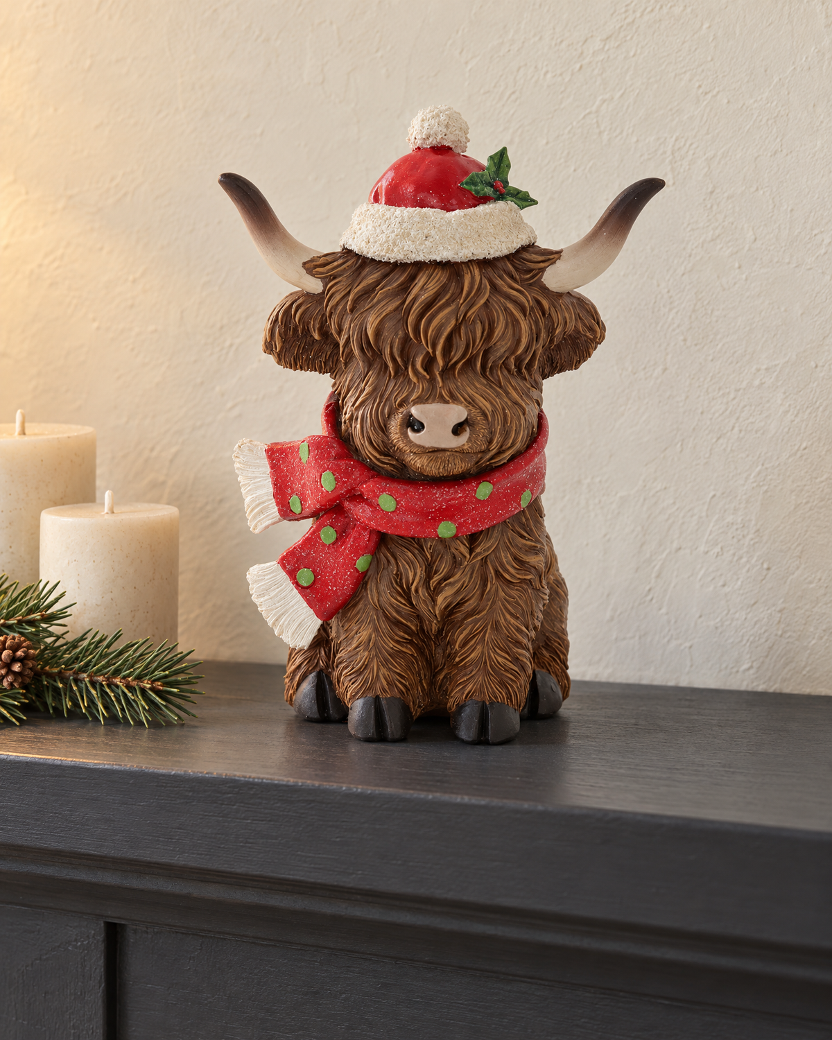 The Highland Cow Christmas figure on a charcoal shelf beside cream candles and a pine sprig