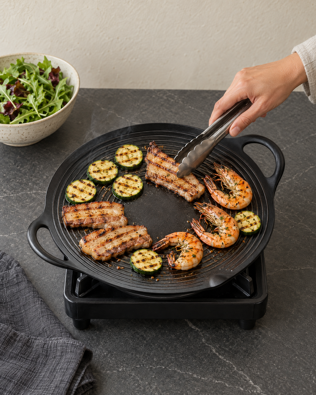 Cast iron grill pan on a portable gas stove with pork belly, prawns and courgette, turned with tongs
