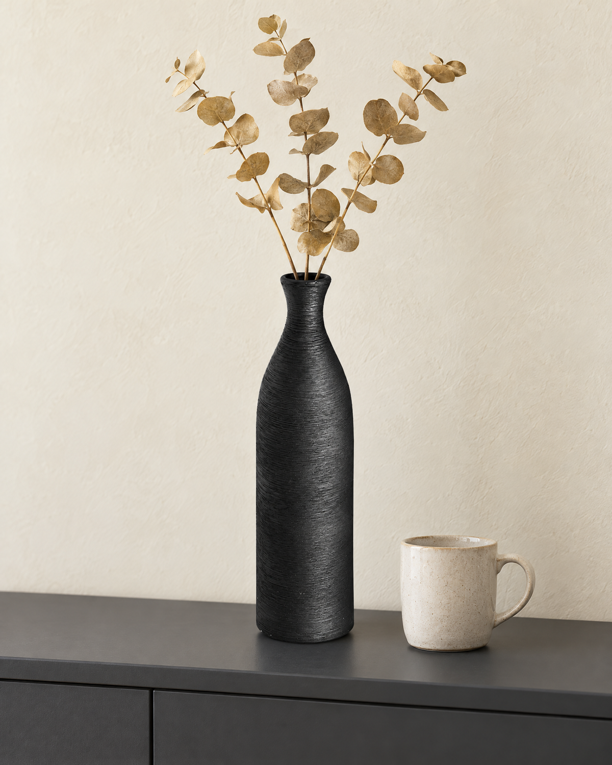 The black bottle vase with dried golden eucalyptus stems on a charcoal sideboard beside a mug