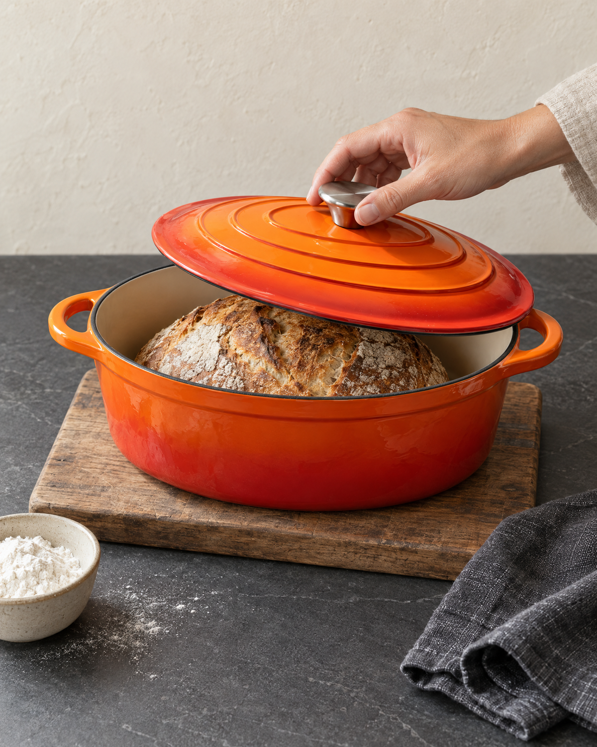 Lid lifted off the orange oval Dutch oven to show a baked sourdough loaf inside, on a wooden board