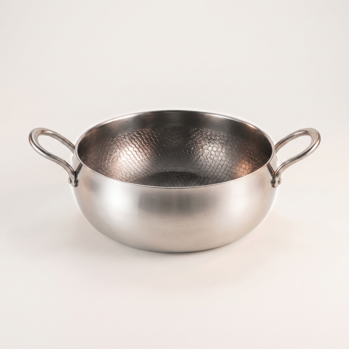 Deep hammered titanium pot with two riveted loop handles on a seamless off-white paper sweep.
