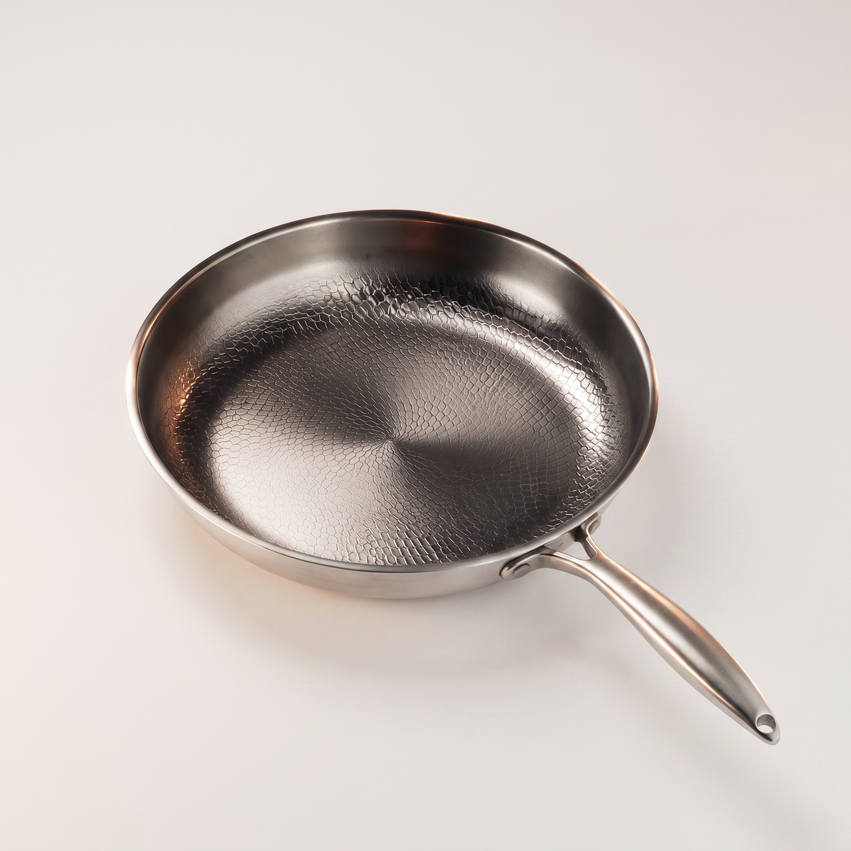 The same pan seen from a high angle alone on a plain light grey backdrop, its riveted handle running out to the lower right and ending in a hanging hole.