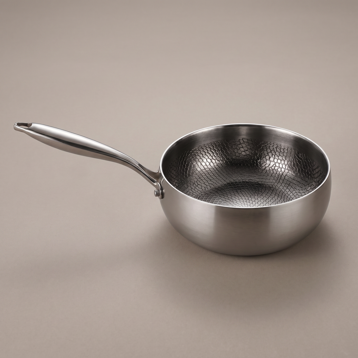 Saute pan standing empty on a plain greige backdrop, its honeycomb textured interior and rounded body lit from above, with a long polished handle ending in a hanging slot.