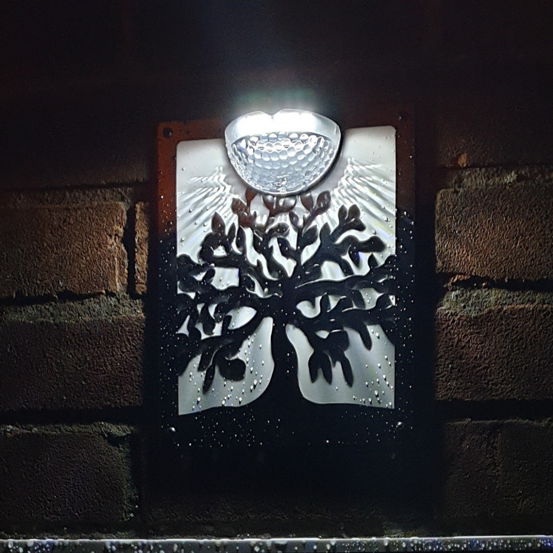 Tree of life solar plaque with white backing panel lit at night on a brick wall